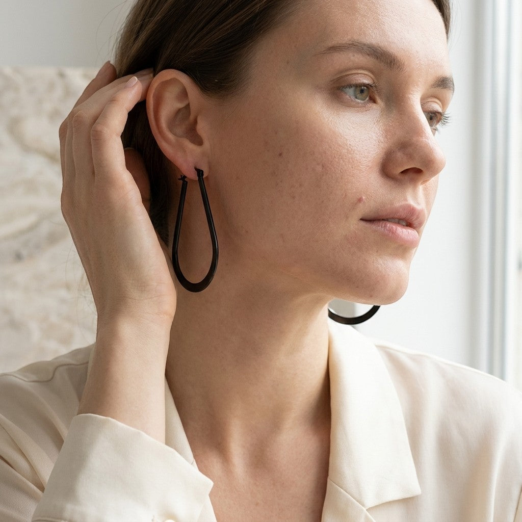 Aramat Jewels black oval hoop earrings worn by a woman with her hand near her ear. The earrings are made of steel and have a shiny finish.