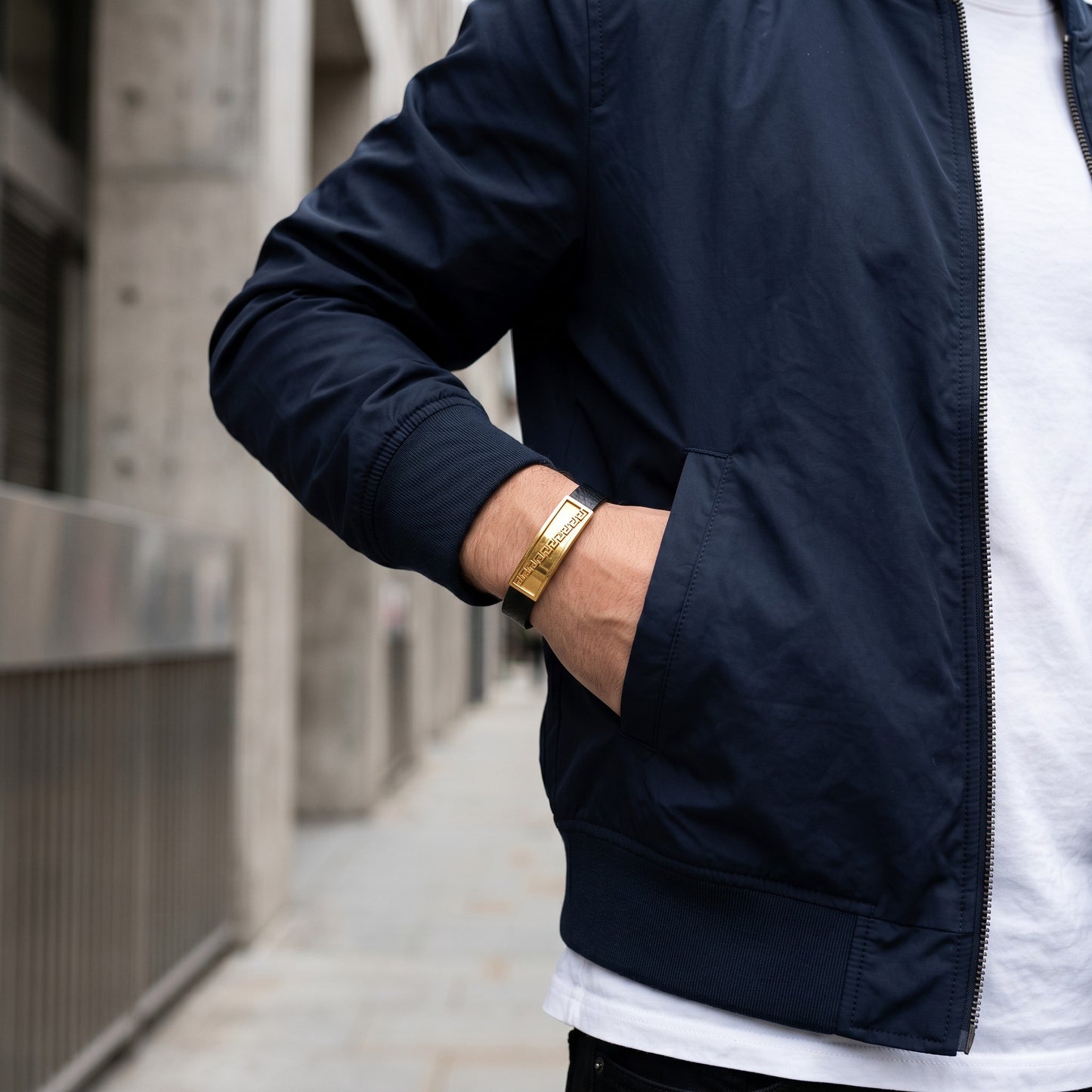 Aramat Jewels bruine leren herenarmband met goudkleurige stalen plaat met Grieks motief gedragen bij een jas