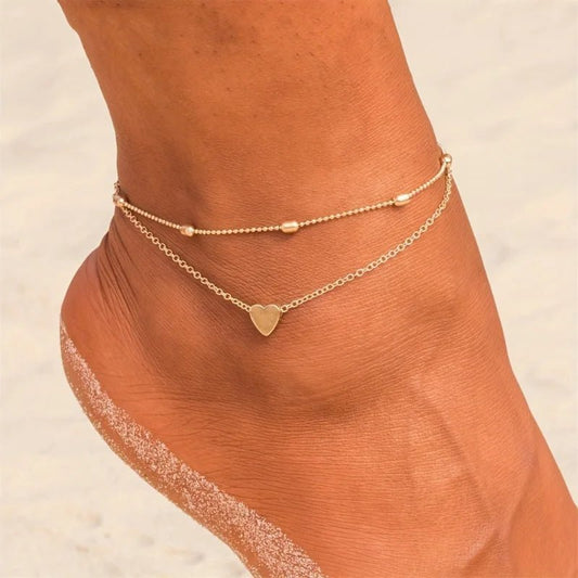 Aramat Jewels gold-tone double-layered steel anklet featuring a small heart charm and delicate beads on a woman's ankle.