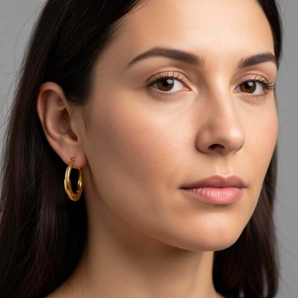 Aramat Jewels small gold-coloured stainless steel hoop earrings, 24x23mm, with a hinge clasp, worn by a woman.