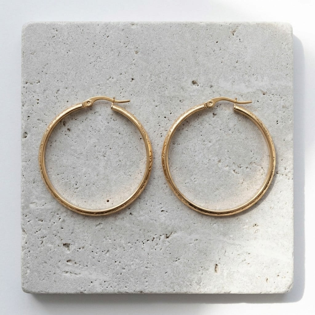 Aramat Jewels gold stainless steel hoop earrings with floral motif, 40mm diameter, on a textured stone surface.