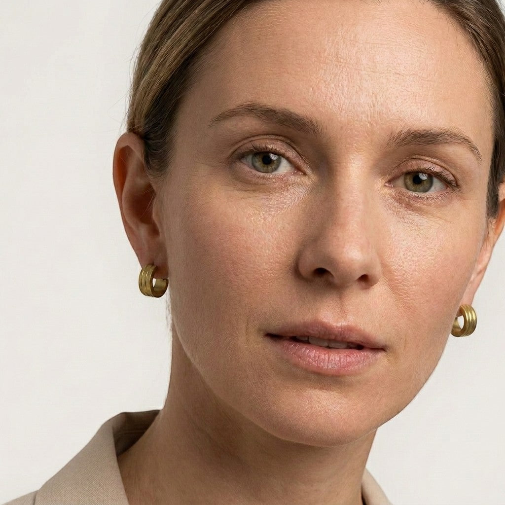 Aramat Jewels gold-tone stainless steel huggie hoop earrings with sandblasted stripes, worn by a woman.