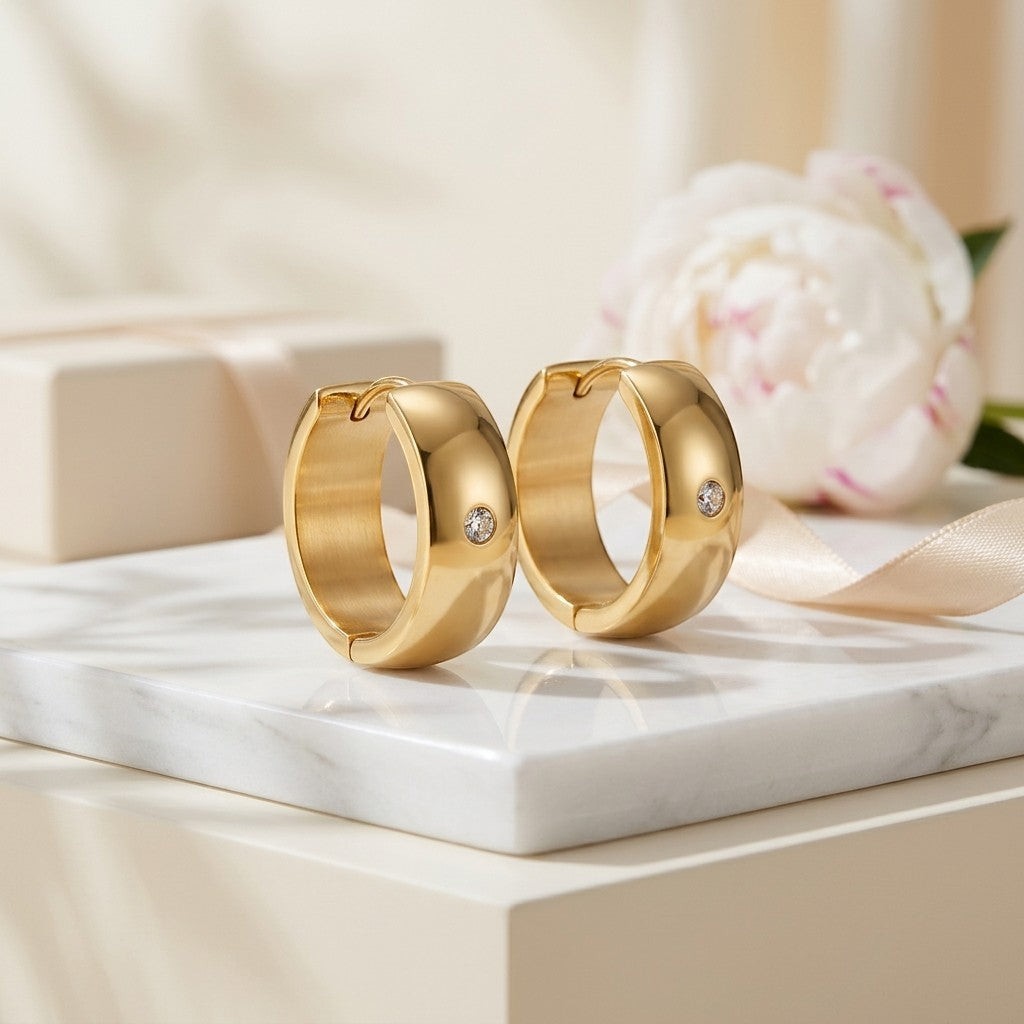 Aramat Jewels gold-tone stainless steel huggie hoop earrings with a central cubic zirconia stone, displayed on a marble surface with a gift box and flower.