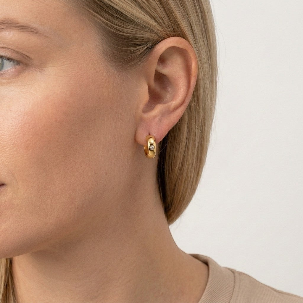 Aramat Jewels gold-tone stainless steel huggie hoop earrings with a central cubic zirconia stone, worn by a model.
