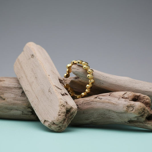 Aramat Jewels gold-colored stainless steel adjustable ring with an oval block pattern, displayed on driftwood.