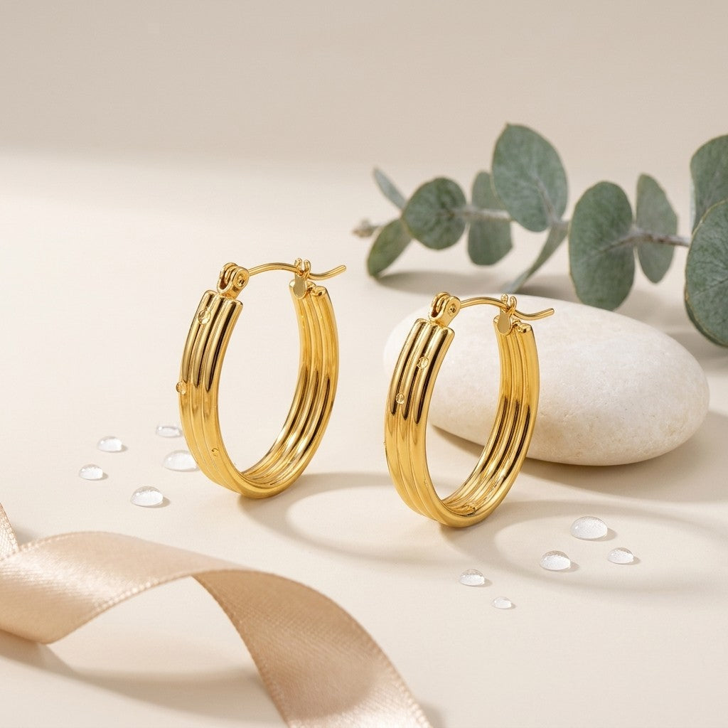 Aramat Jewels gold oval hoop earrings with ribbed texture and small dot details, 26mm. Styled with eucalyptus leaves and a smooth stone.