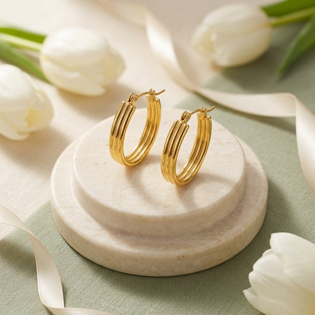 Aramat Jewels gold oval hoop earrings with a ribbed design, displayed on a marble stand with white tulips and ribbon.