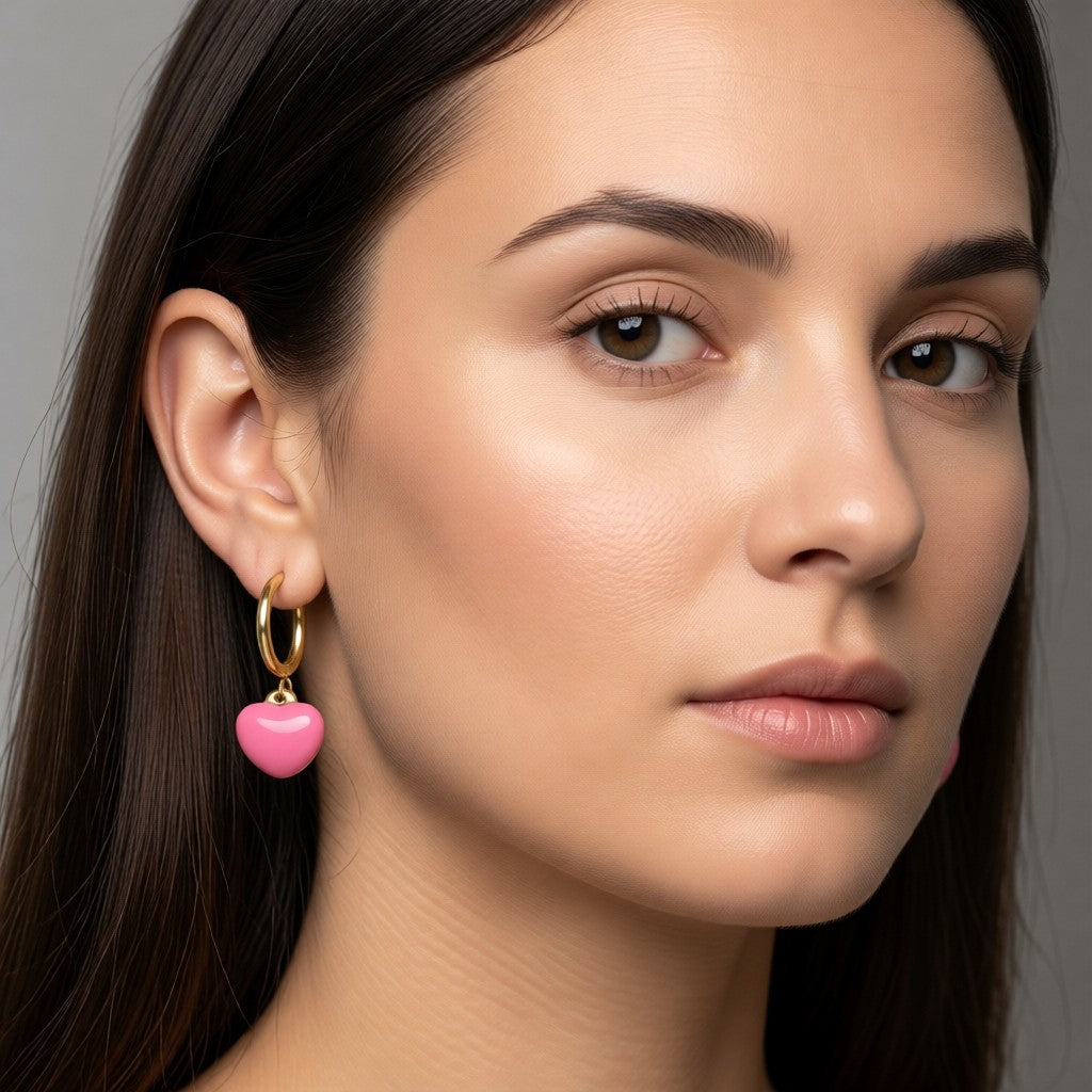 Aramat Jewels gold huggie hoop earrings with a pink enamel heart charm, worn by a woman.