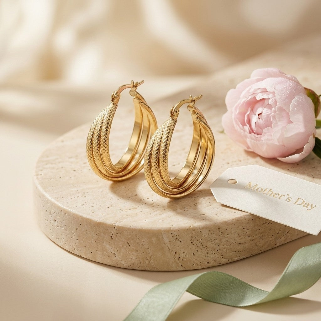Aramat Jewels gold triple hoop earrings with textured stripe pattern, 25mm diameter, displayed on a stone pedestal with a pink peony and Mother's Day tag.