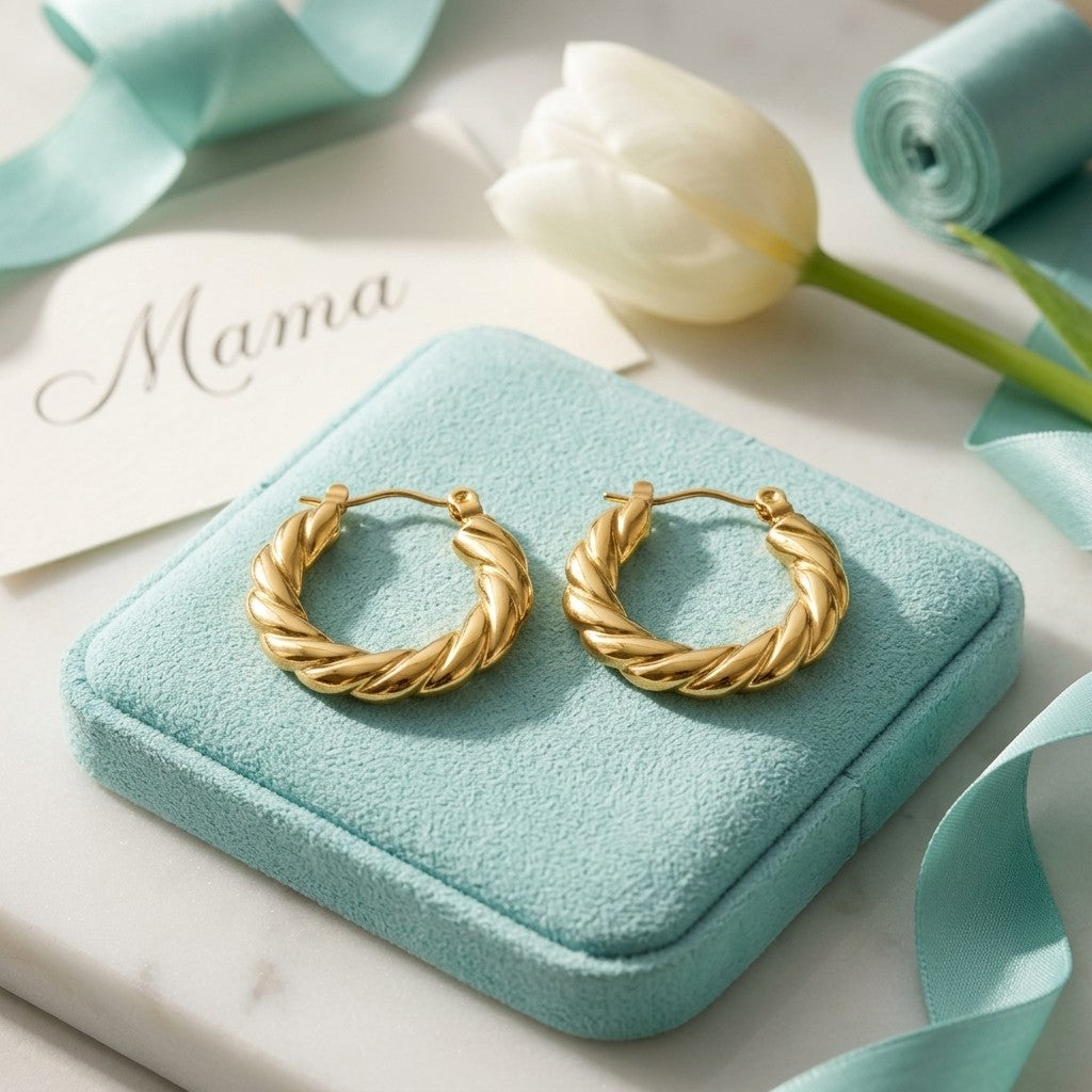 Aramat Jewels gold-tone stainless steel twisted hoop earrings, 23mm diameter, displayed on a light blue velvet stand.