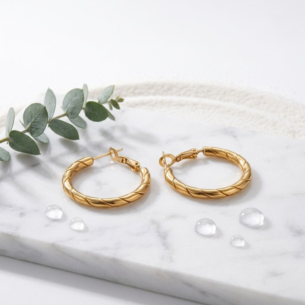 Aramat Jewels gold-tone stainless steel hoop earrings with a twisted rope motif, 25x3.5mm, on a marble surface with water droplets and eucalyptus leaves.