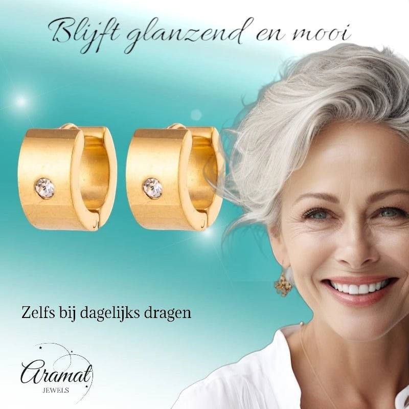 Aramat Jewels gold-colored wide stainless steel huggie hoop earrings with a single clear zirconia stone, worn by a smiling woman with grey hair.