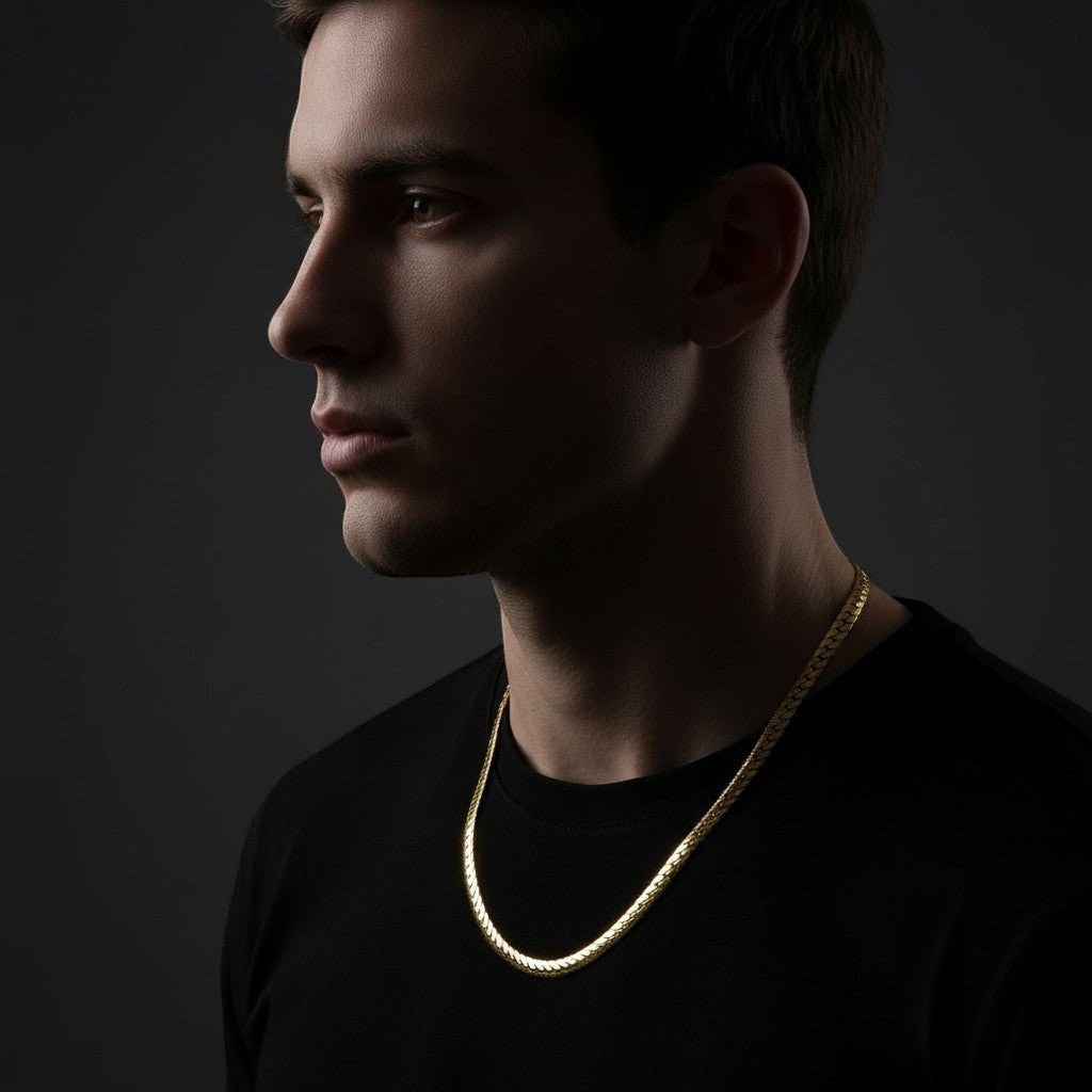 Man draagt een 6mm goudkleurige Aramat Jewels gourmet ketting van staal met een bewerkte rand tegen een donkere achtergrond