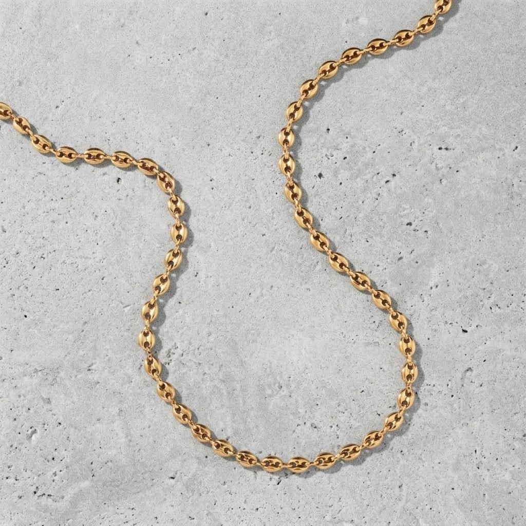 Aramat Jewels goudkleurige koffieboon ketting van 316L staal met 4,5mm schakels op een grijze betonnen achtergrond.