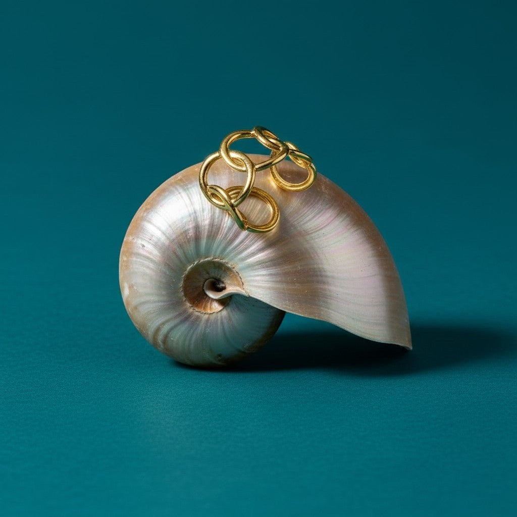 Aramat Jewels goudkleurige RVS ring met ovale schakels, gepresenteerd op een schelp tegen een blauwgroene achtergrond.