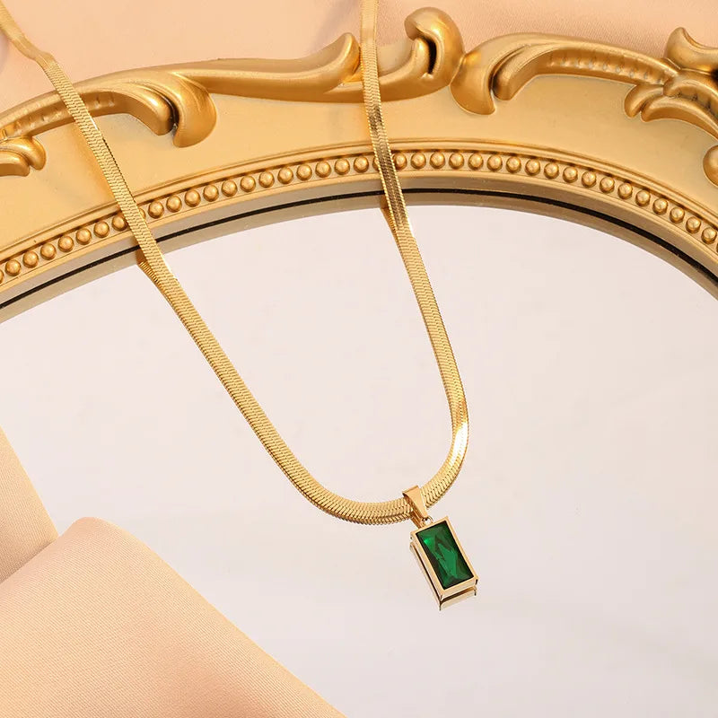 Aramat Jewels goudkleurige platte slangenketting met een rechthoekige groene zirkonia hanger op een spiegel.