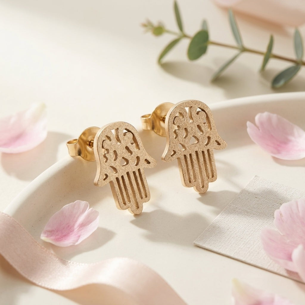 Aramat Jewels goudkleurige Hamsa oorstekers van RVS met een matte, gezandstraalde afwerking op een sierlijk schaaltje.