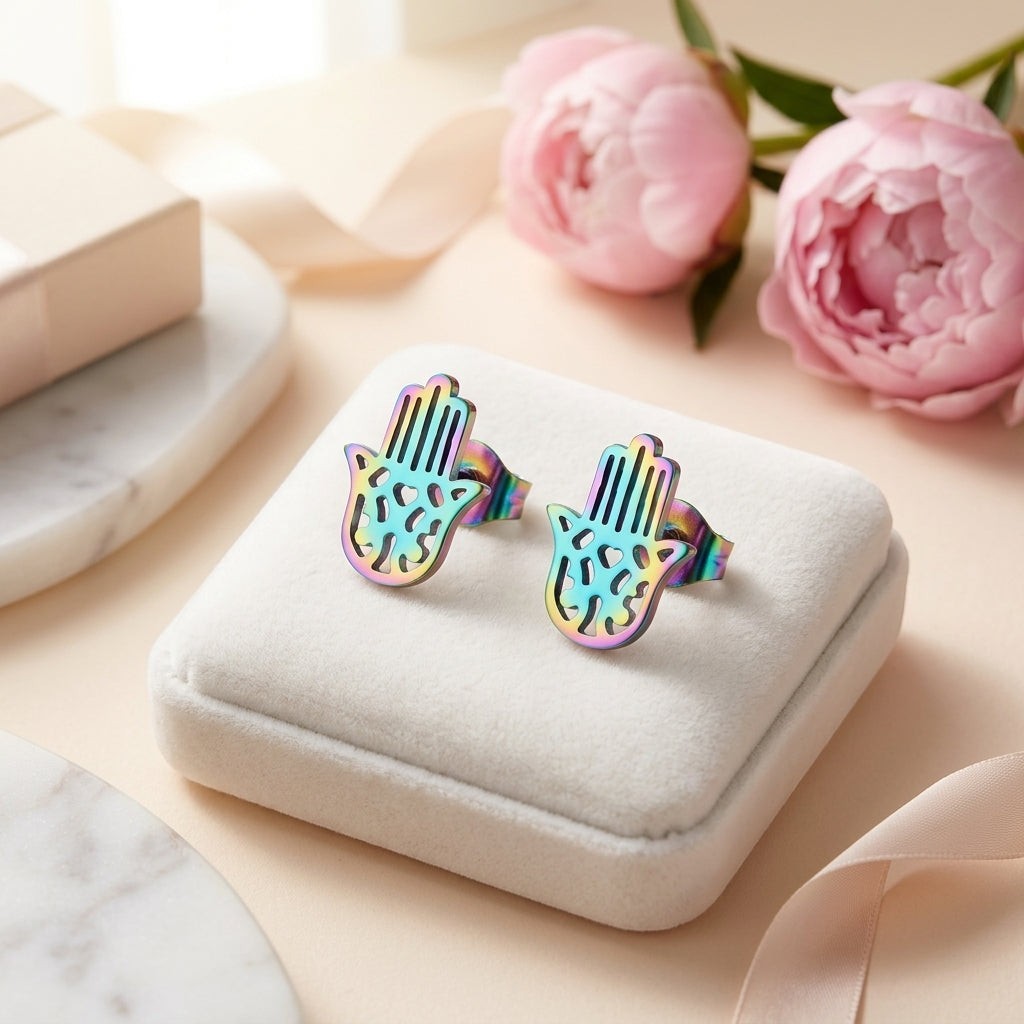 Aramat Jewels multikleur Hamsa oorstekers van RVS met regenboogglans op een wit sieradendoosje naast roze bloemen.