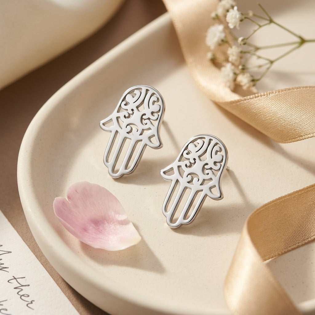 Aramat Jewels zilverkleurige Hamsa hand oorknopjes van RVS met gedetailleerd filigraan patroon op een beige schaaltje.
