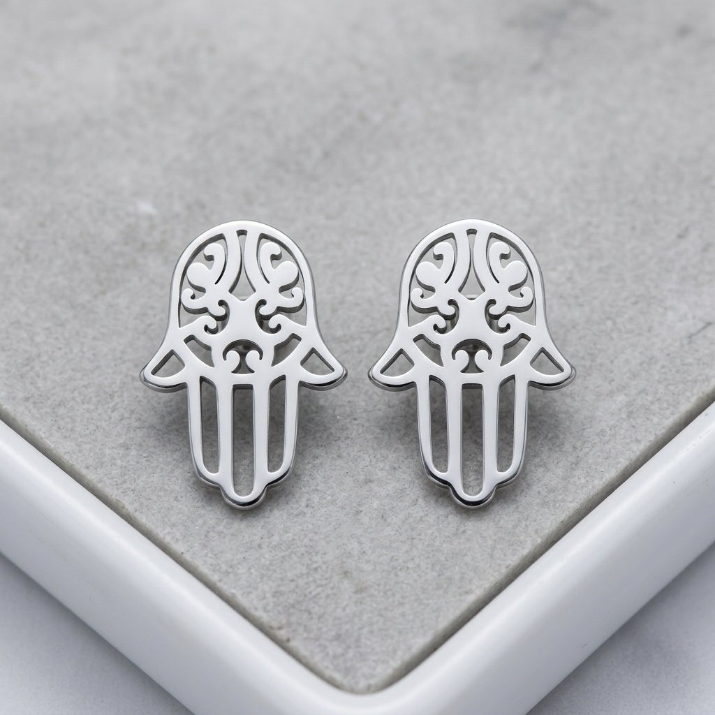 Aramat Jewels zilverkleurige Hamsa hand oorknopjes van RVS met een glanzende afwerking op een grijze ondergrond.