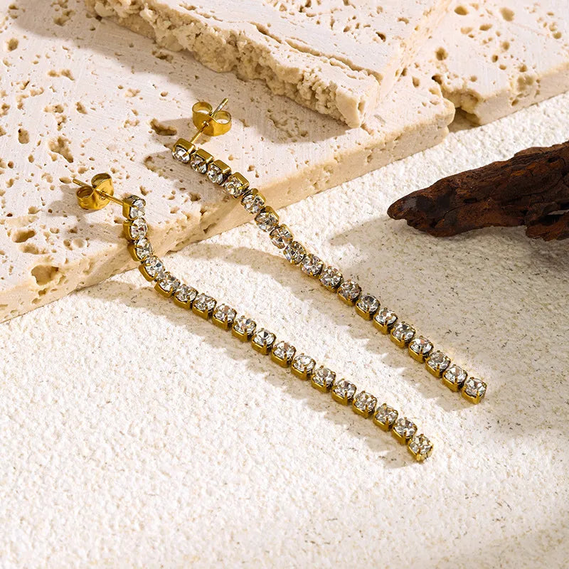 Aramat Jewels lange goudkleurige RVS oorhangers met transparante kristallen op een gestructureerde ondergrond.