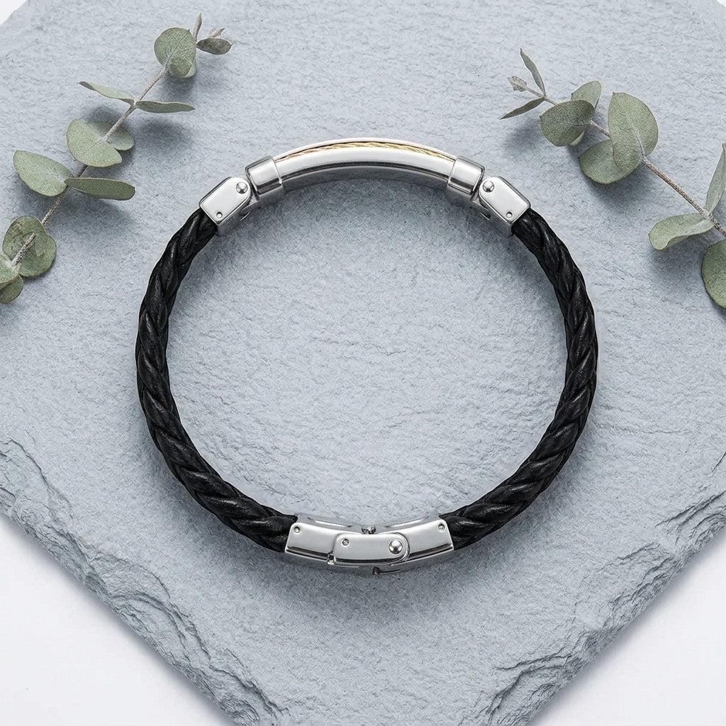 Aramat Jewels zwarte gevlochten leren armband met roestvrijstalen accenten in zilver, goud en rosé op een stenen plaat.