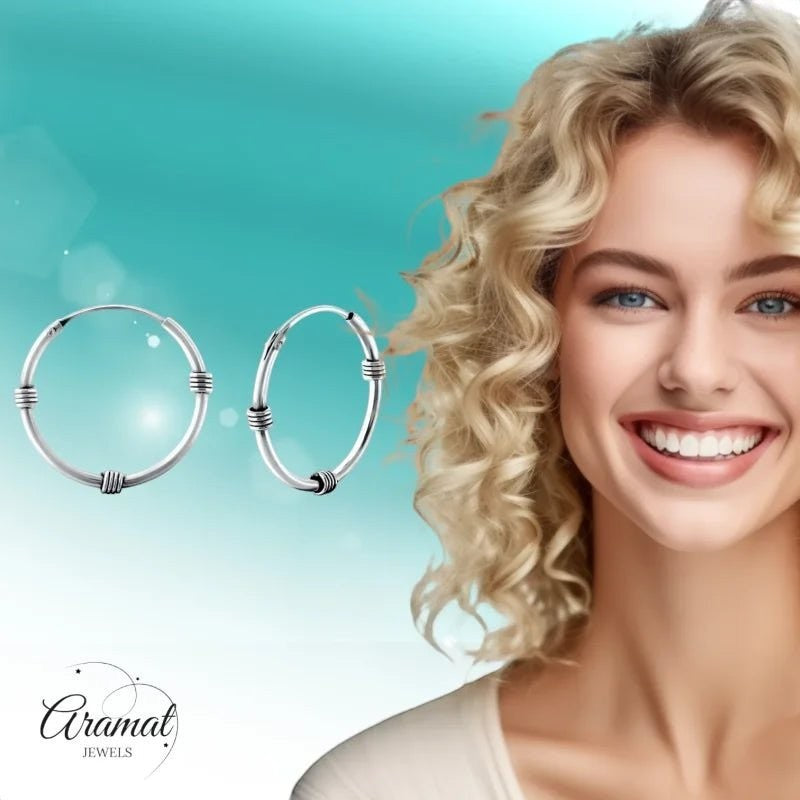 Aramat Jewels 20mm oxidized silver Bali hoop earrings with textured detailing. A smiling woman with blonde curly hair is partially visible on the right.
