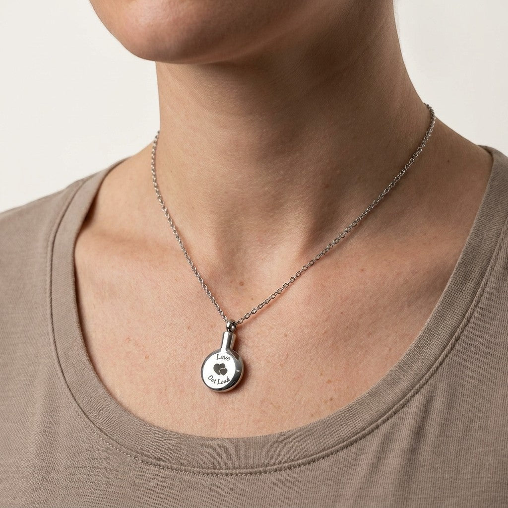 Aramat Jewels zilverkleurige ronde ashanger met gravure 'love out loud' en twee hartjes, gedragen door een vrouw.