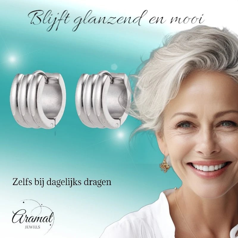 Aramat Jewels wide silver-tone stainless steel hoop earrings with ribbed detail, worn by a smiling woman with grey hair.