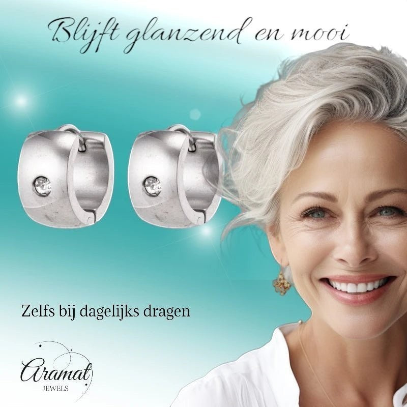 Aramat Jewels silver-colored wide stainless steel hoop earrings with a clear zirconia stone, worn by a smiling woman with grey hair.