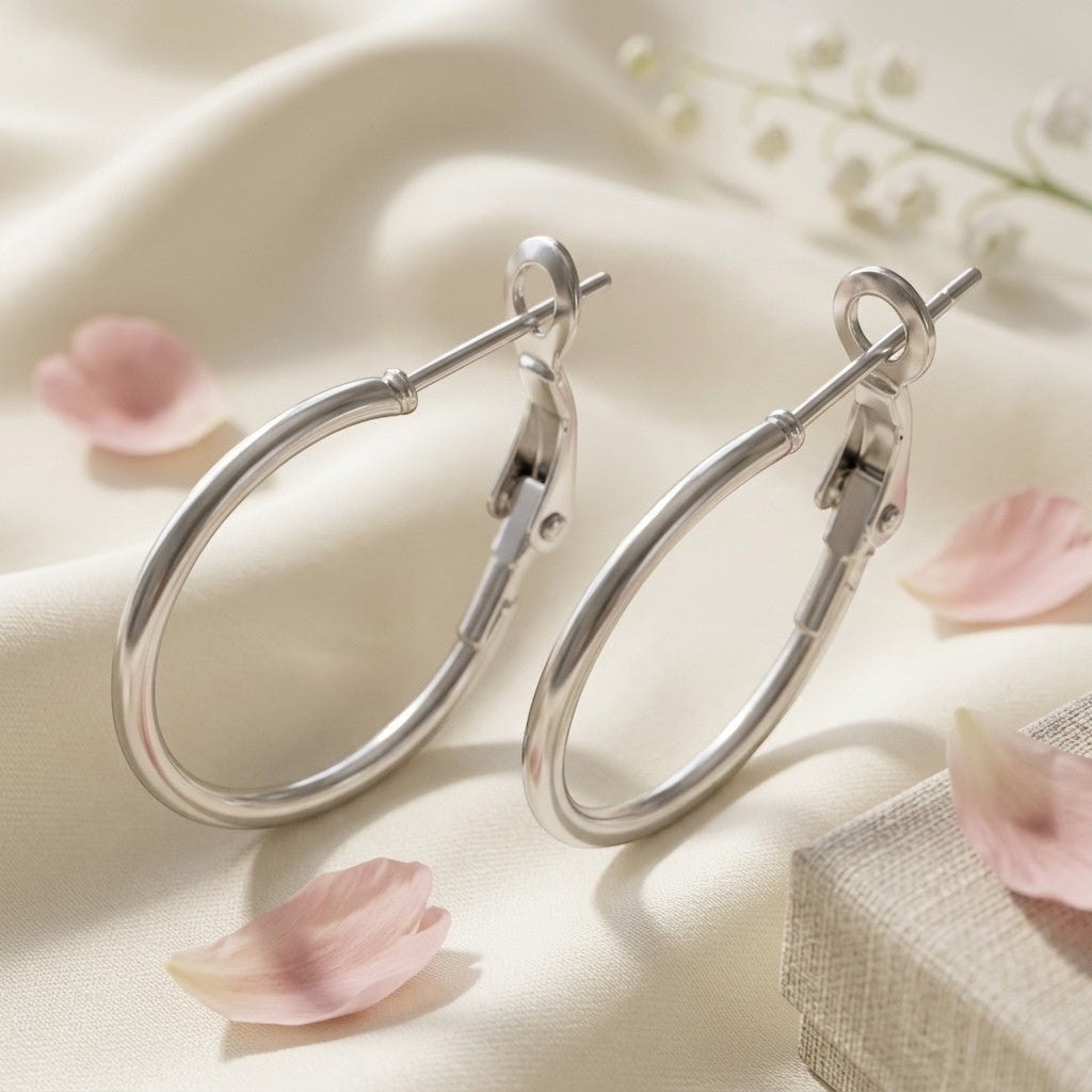 Aramat Jewels silver-tone stainless steel hoop earrings with a high-gloss finish and hinge clasp, shown on a cream silk background with pink petals.