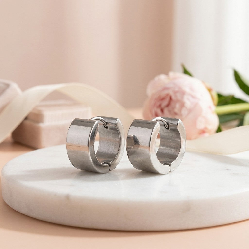Aramat Jewels wide silver stainless steel huggie hoop earrings with a smooth finish, displayed on a marble surface with soft pink background elements.