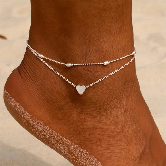 Aramat Jewels silver-colored steel anklet set featuring a delicate heart charm and small beads on a beach background.