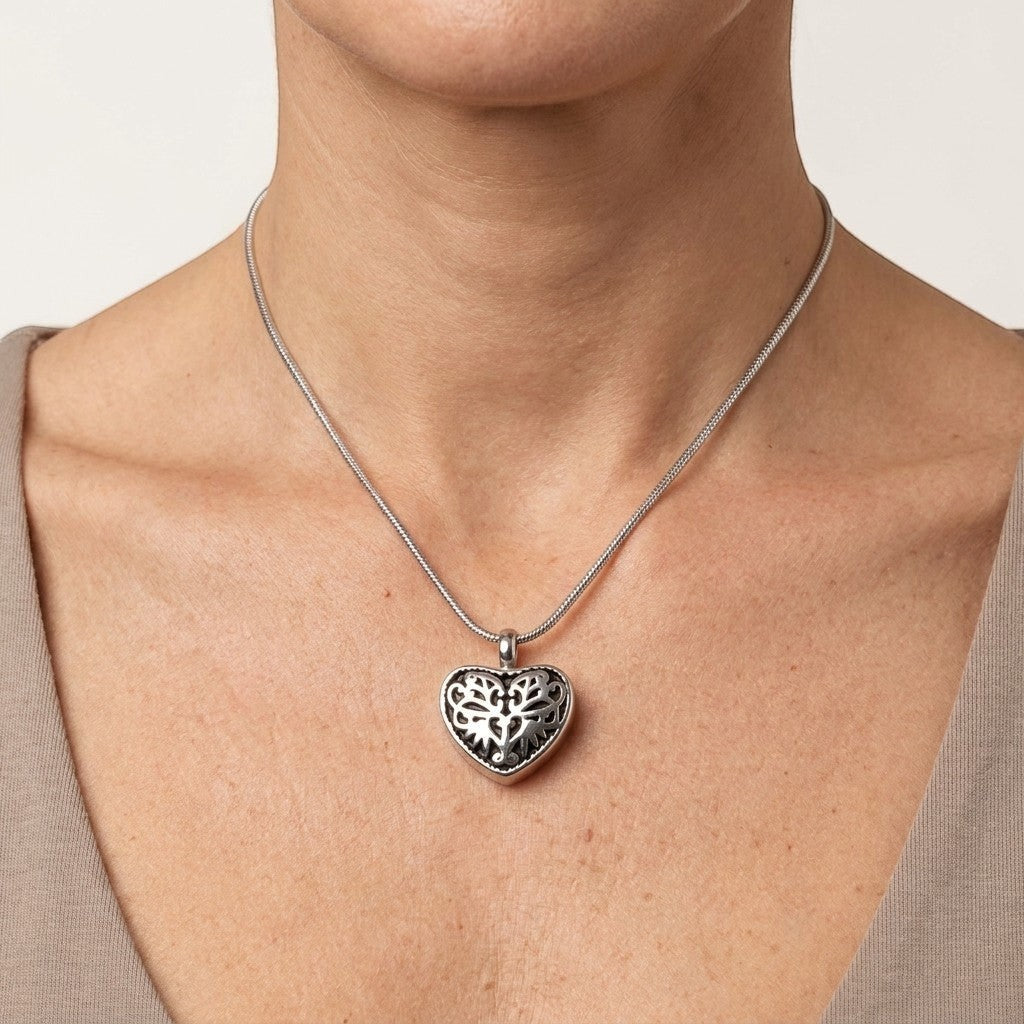 Aramat Jewels silver-tone steel heart-shaped cremation pendant with black enamel inlay and intricate filigree design, worn on a snake chain necklace.