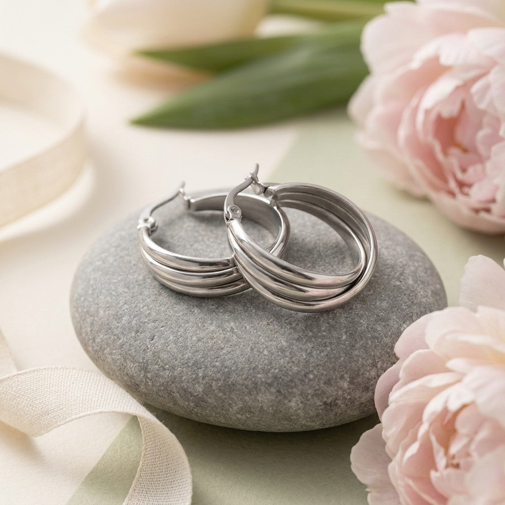 Aramat Jewels silver-tone stainless steel triple hoop earrings with a crossed design, 20x7.5mm, on a grey stone with soft pink flowers.