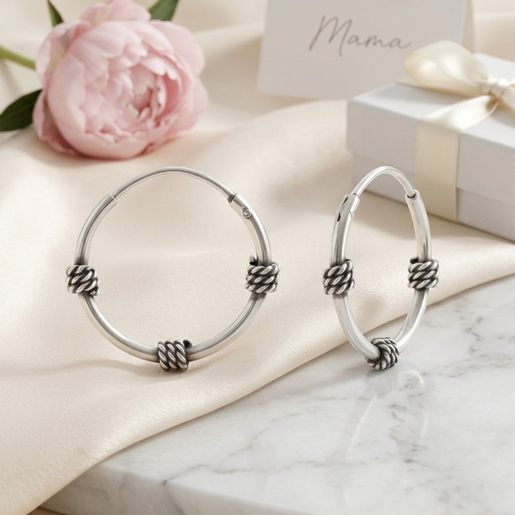 Aramat Jewels thin 16mm silver Bali hoop earrings with a braided detail, presented on a satin cloth with a pink peony and gift box.