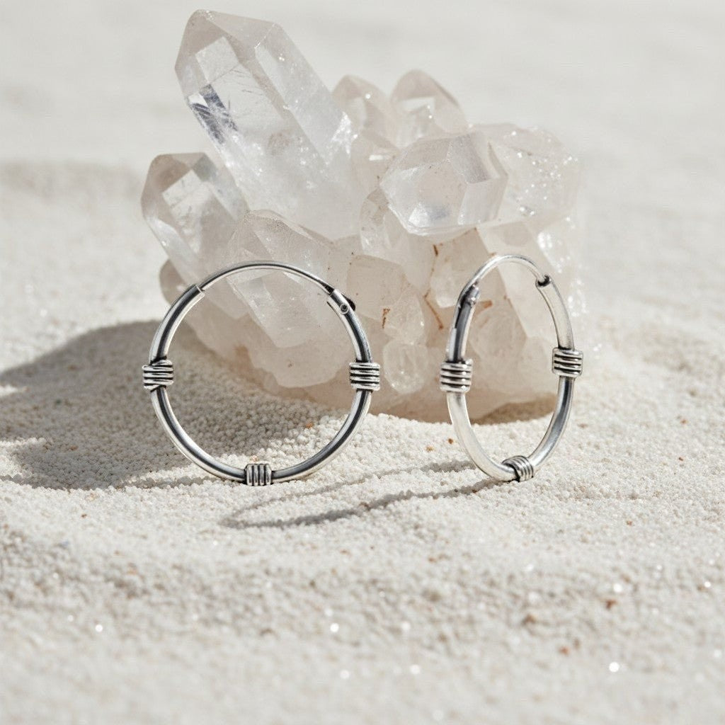 Aramat Jewels unisex silver Bali hoop earrings, 16mm diameter, with a textured band design, displayed on a sandy surface with crystals.