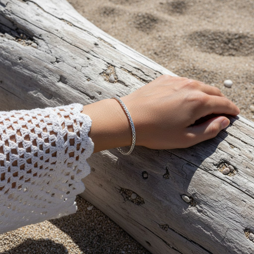 Aramat Jewels zilverkleurige vossenstaart armband van RVS gedragen door een persoon op een strand bij een boomstam.