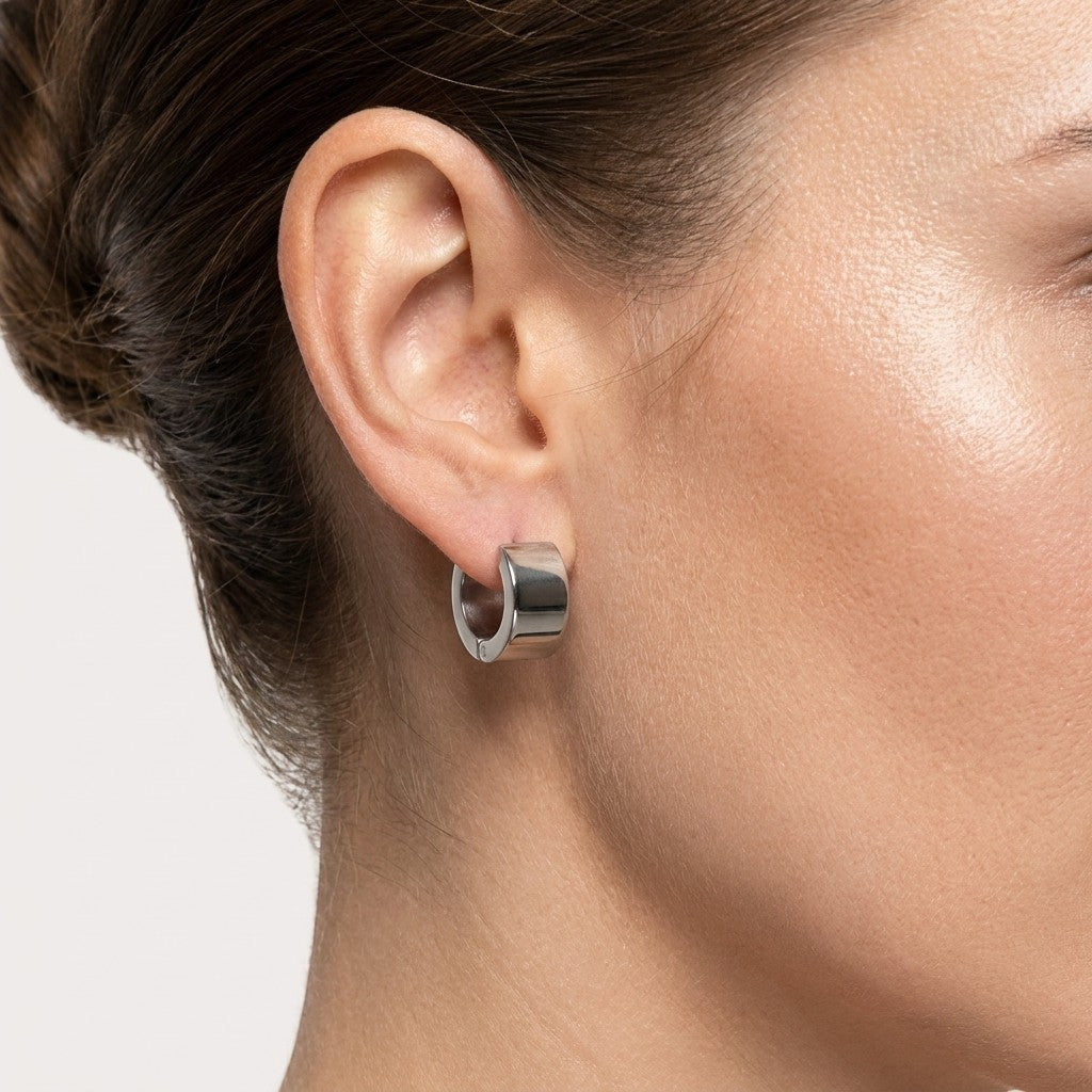 Aramat Jewels wide silver-tone stainless steel hoop earrings on a model's ear.