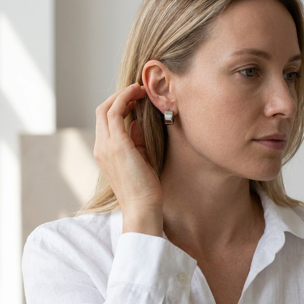 Aramat Jewels wide silver-tone stainless steel huggie hoop earrings worn by a woman with blonde hair.
