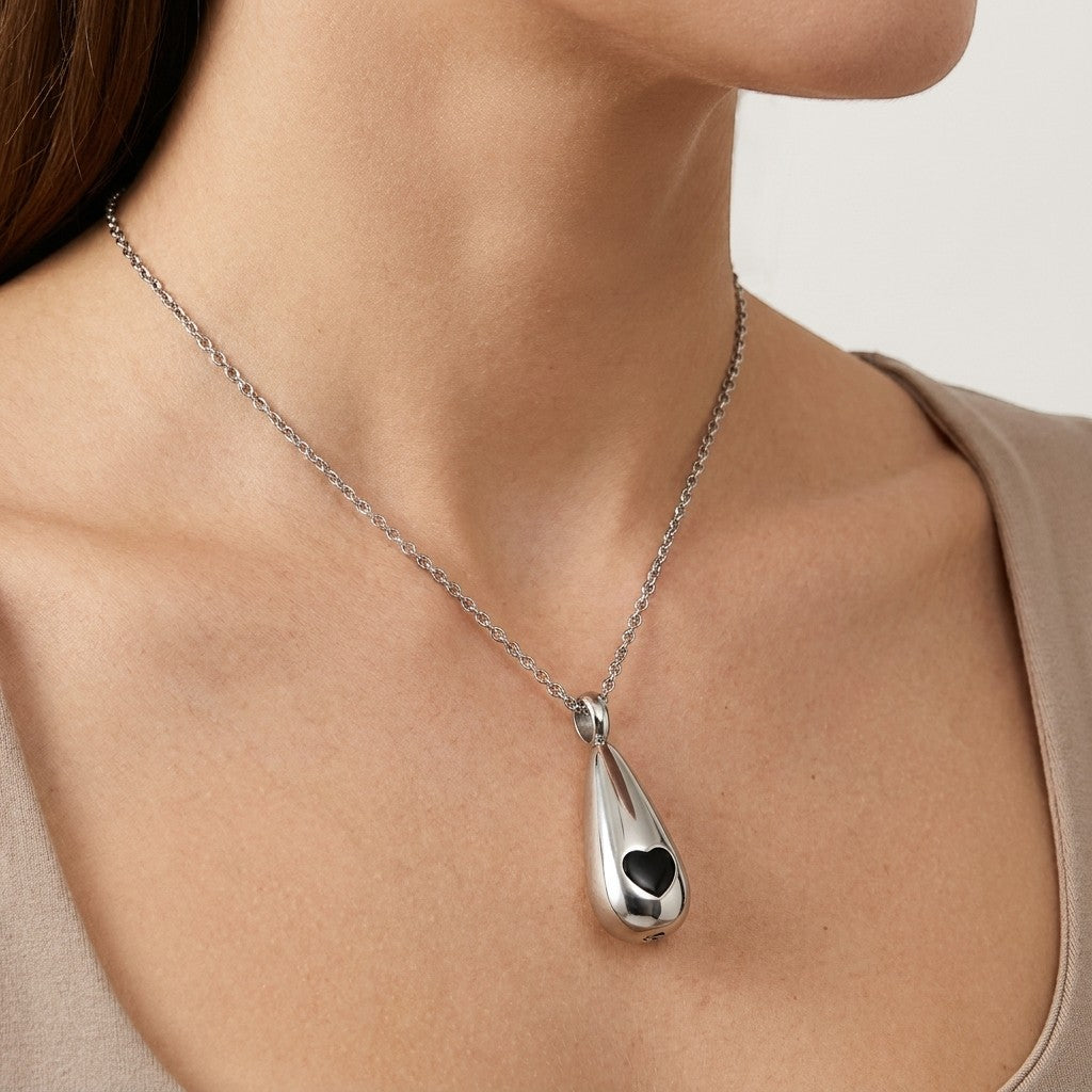 Aramat Jewels zilverkleurige druppel ashanger met zwart hart detail, gemaakt van roestvrij staal.