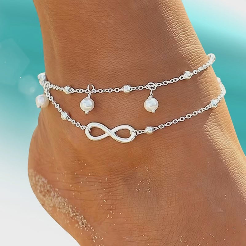 Aramat Jewels dubbel zilverkleurig enkelbandje met infinity teken en parels om een enkel op een strandachtergrond