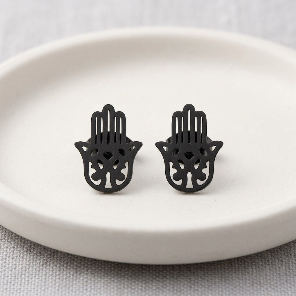 Zwarte Aramat Jewels Hamsa oorstekers van RVS in de vorm van een Handje van Fatima op een wit schaaltje.
