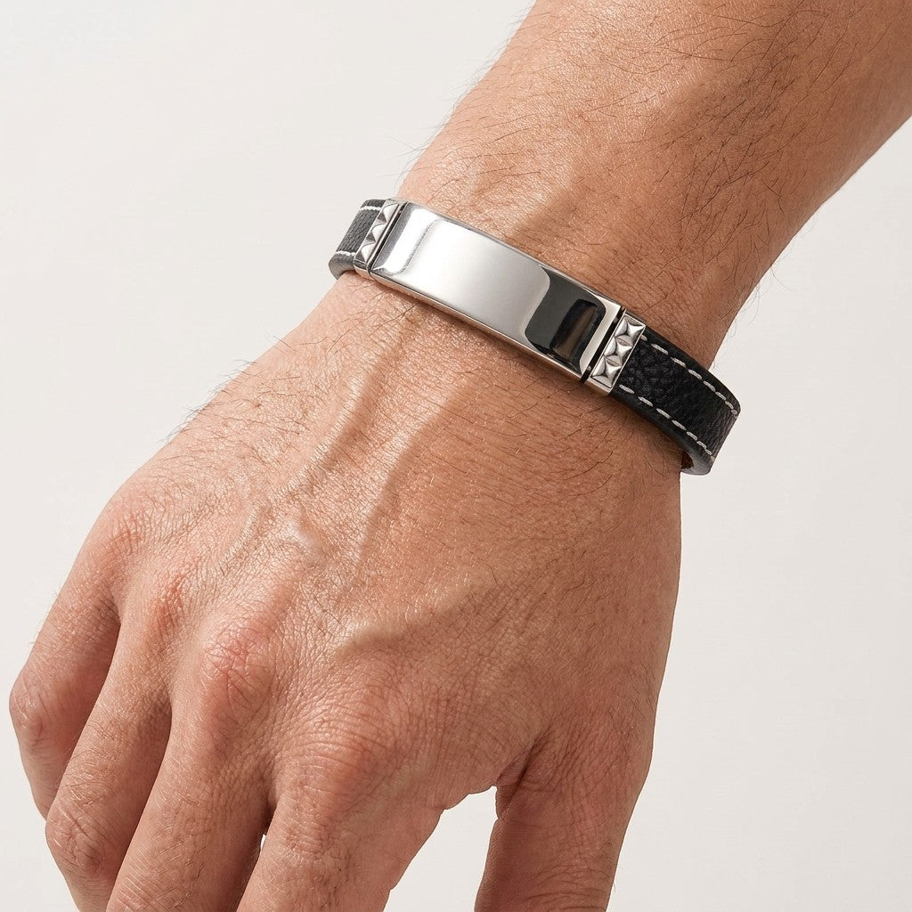 Aramat Jewels zwarte leren herenarmband met graveerbare zilverkleurige 316L RVS plaat en wit stiksel om een pols.