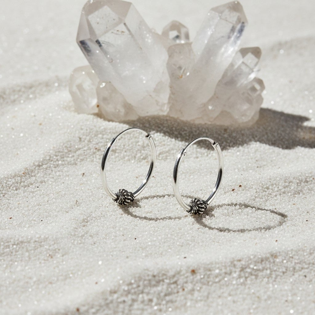 Aramat Jewels 18mm silver Bali hoop earrings with intricate ethnic design, set on white sand with crystals.