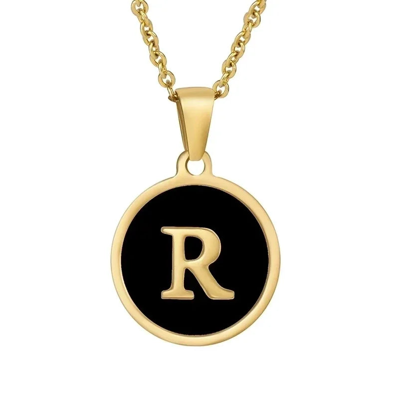 Gouden hangerketting met de letter ’R’ op een zwarte ronde achtergrond.