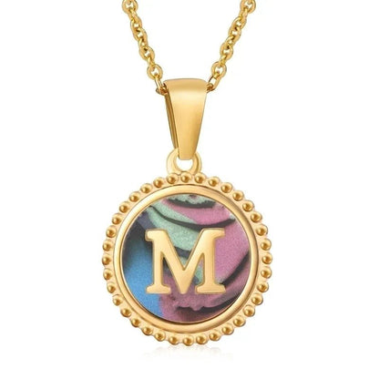 Hanger met vergulde ketting met de letter ’M’ op een kleurrijke iriserende achtergrond met kralenranddetail.