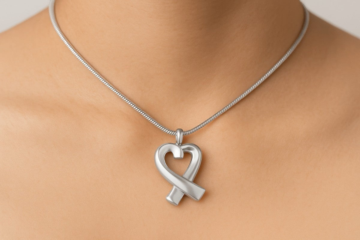 Zilveren hartvormige ashanger met lint-ontwerp, 30x17mm, gedragen op vrouwenhals met zilveren slangenketting. Hoogwaardige gedenksieraad voor asresten, rouwsieraad in hartvorm, elegante ashanger zilver.