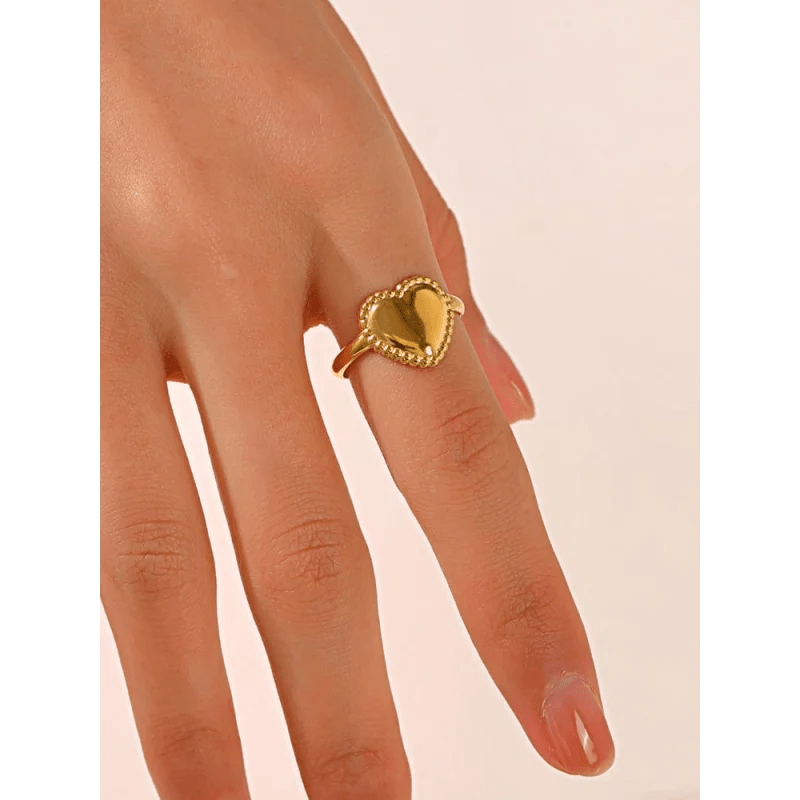 Gouden hartvormige ring met decoratieve rand in touwstijl, gedragen aan een vinger.